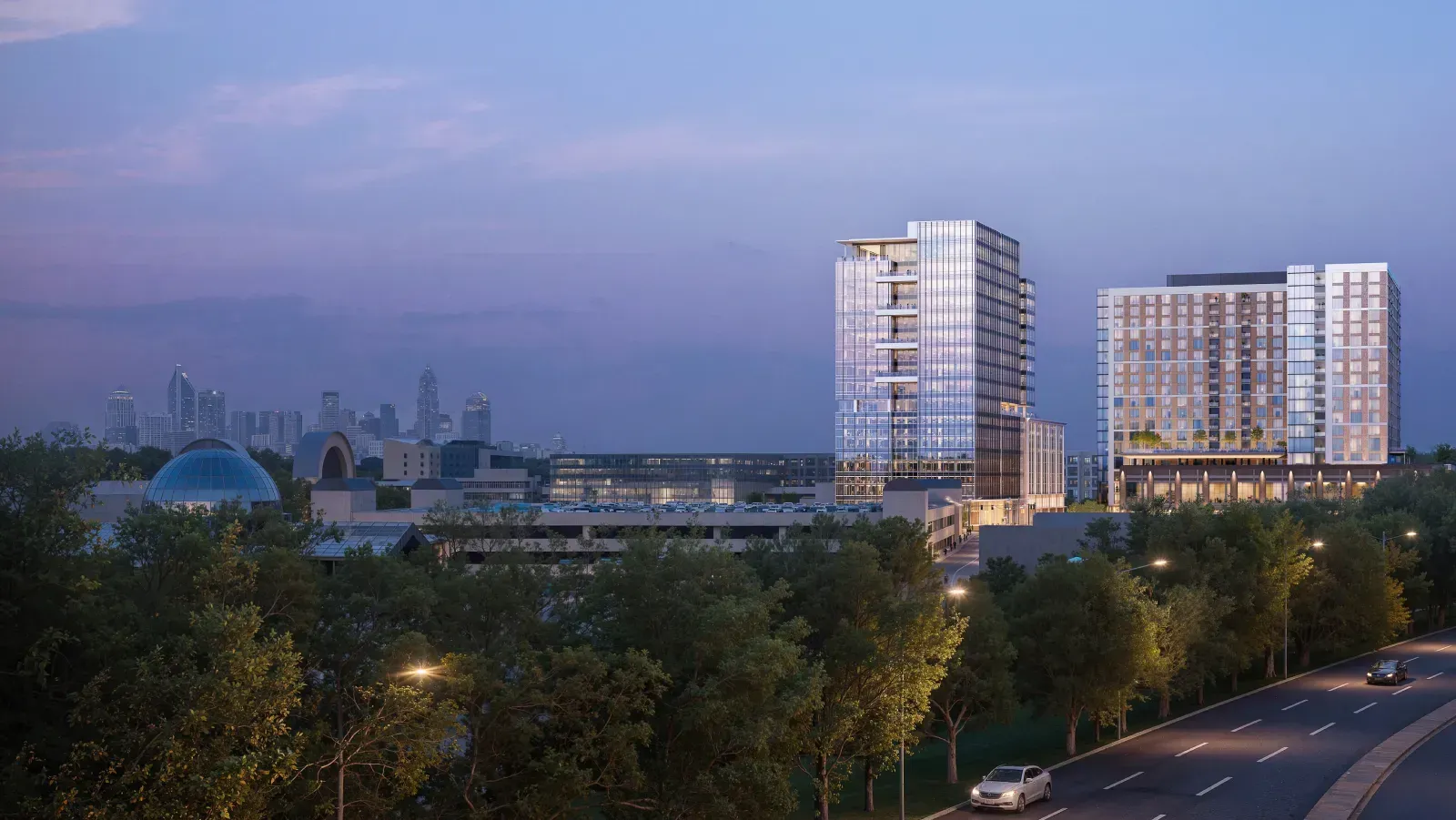 Wide evening architectural rendering showing illuminated high-rise buildings of "The Gallery" complex against a twilight sky, with a city skyline visible in the background to the left.
